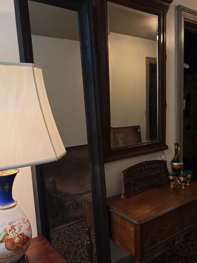 Tall full-length mirror in a dark mahogany frame, leaning against a wall beside a French porcelain table lamp.