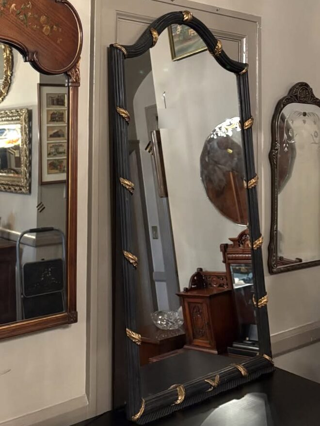 Tall arched mirror in a black reeded frame bound at intervals with gilt leaf wraps, standing on a black table.