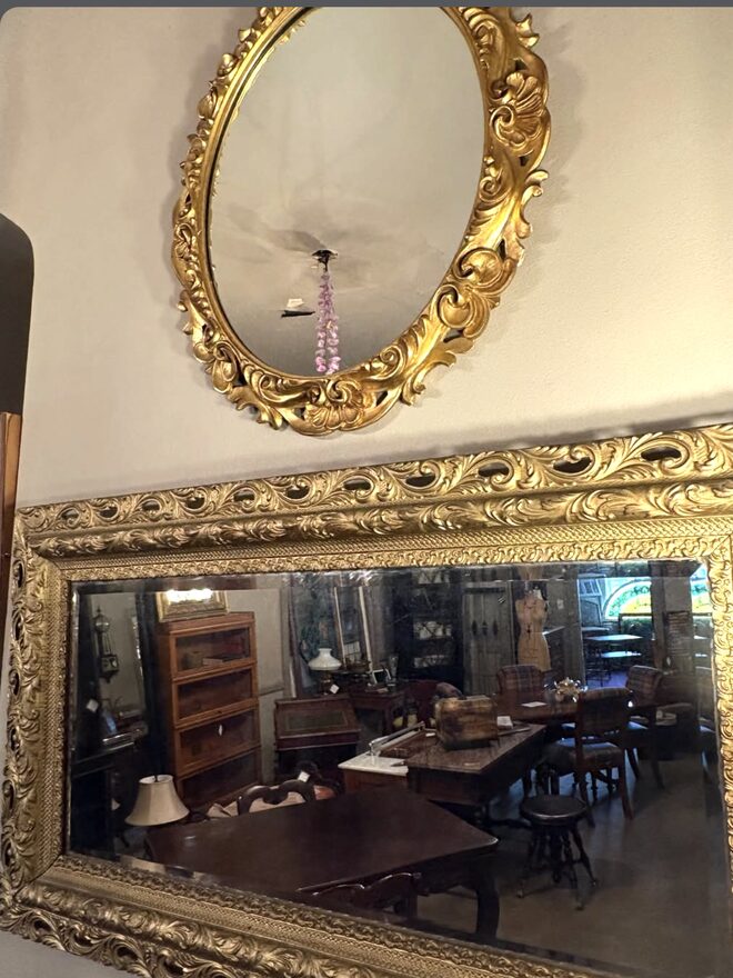 Two gilt mirrors on a wall: an oval mirror in a pierced shell-and-scroll frame hung above a long rectangular beveled mirror in a deep frame of pierced acanthus scrolls.