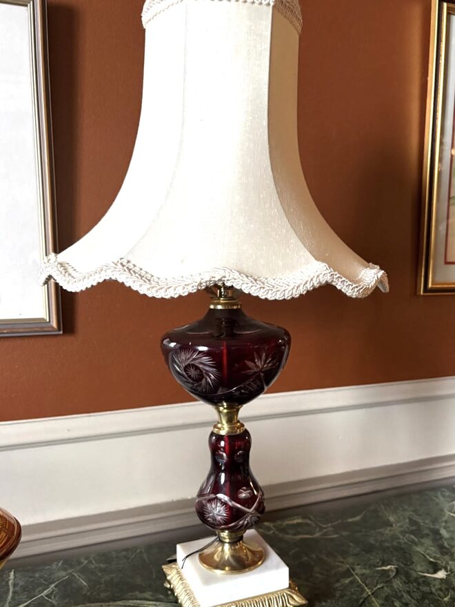 Table lamp with a ruby-red glass font and baluster stem cut through to clear in star and pinwheel patterns, on a white marble plinth and a footed gilt-brass base, under a scalloped cream shade with braided trim.