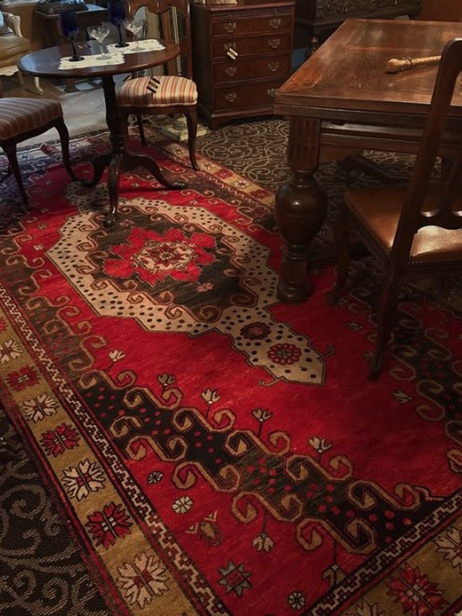 Hand-knotted wool area rug with a deep red ground and a large central medallion, on the floor among antique tables and chairs.