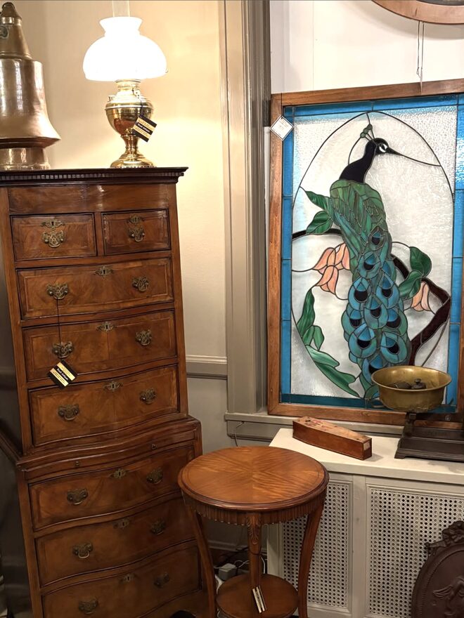 Tall figured-walnut chest-on-chest with a dentil cornice, two short drawers over three long ones above, three long serpentine drawers below, crossbanded drawer fronts and pierced brass pulls, beside a stained-glass peacock panel.